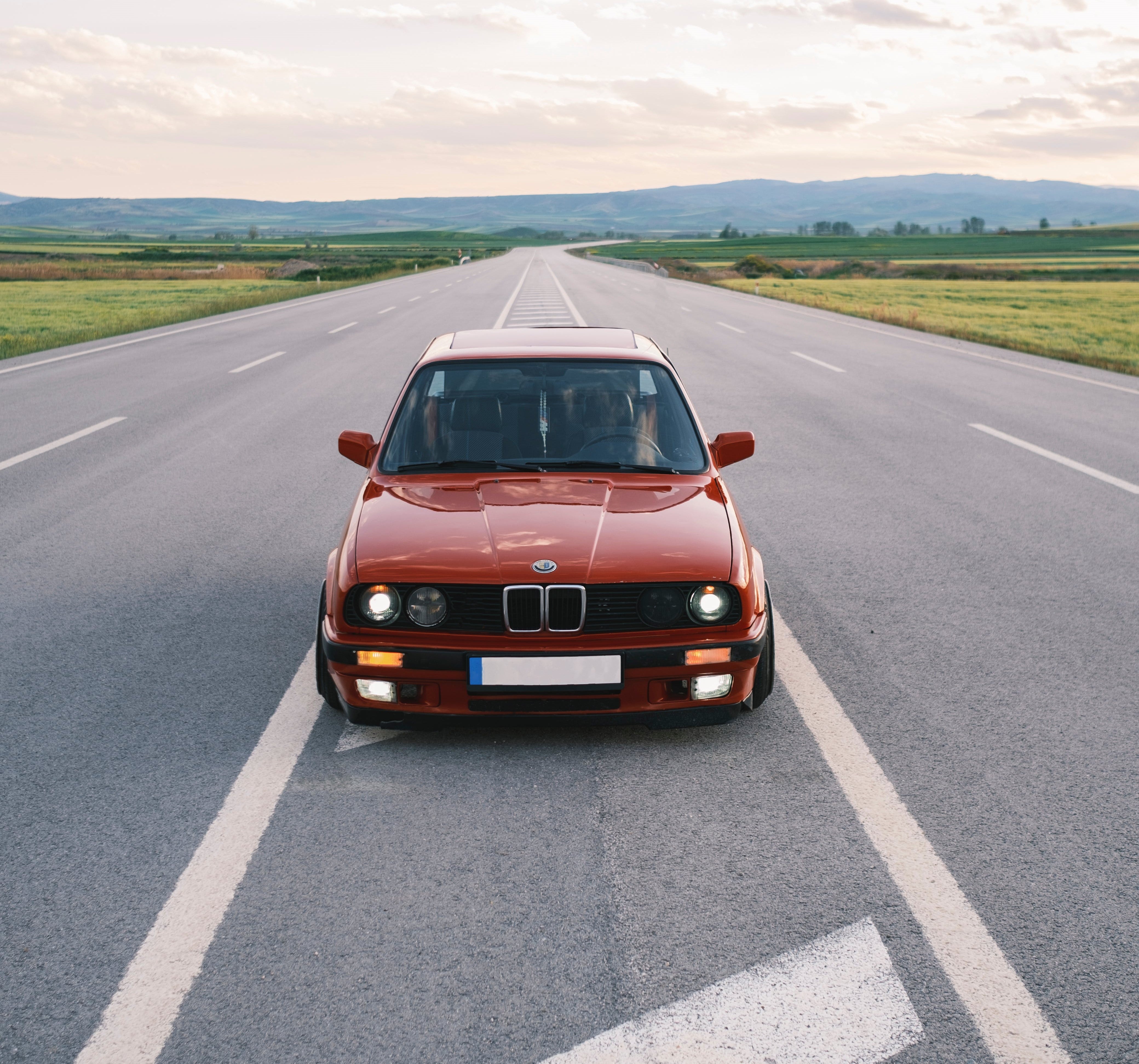 Front view of a red BMW car on a wide, open road surrounded by green fields and mountains.